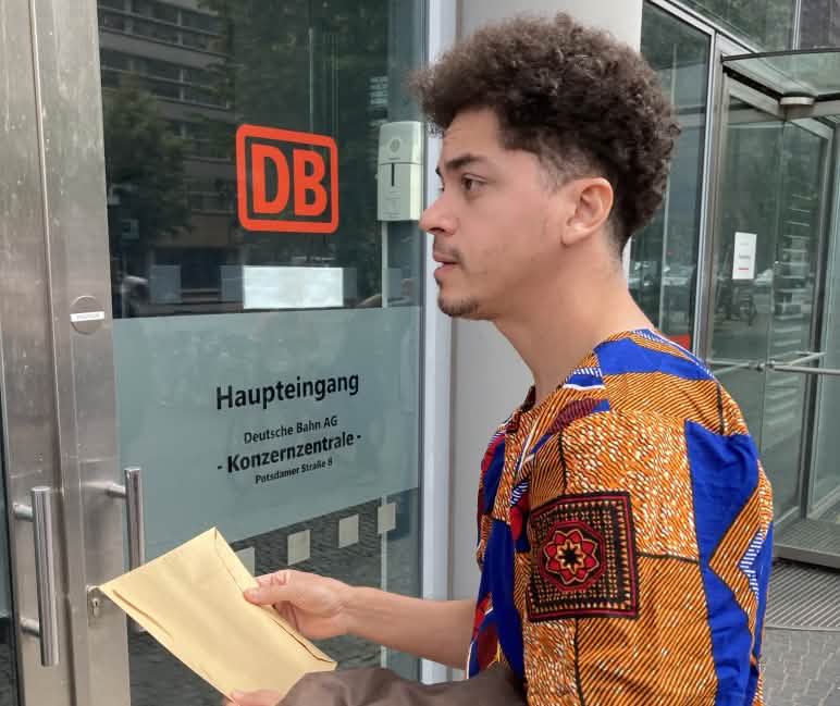 Ein junger Mann steht mit einem Briefumschlag vor einer Glastür mit der Aufschrift DB Haupteingang Konzernzentrale