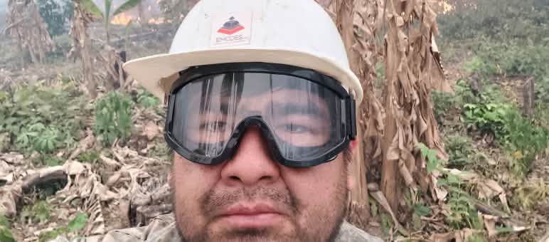 Julio Yeguaori, einer der besten Trompeter Boliviens und Lehrer und Dirigent des Sinfonieorchesters in Urubicha, hilft bei den Löscharbeiten Ein Mann blickt mit einem Helm und einer Schutzbrille in die Kamera, während im Hintergrund Büsche brennen und Rauch aufsteigt