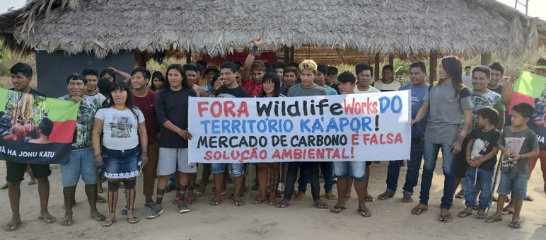 Protest der Ka'apor gegen Kohlenstoffhändler in ihrem Territorium. Aufschrift auf dem Banner: Wildlife Works raus aus dem Ka'apor-Territorium! Der Kohlenstoffmarkt ist eine falsche Umweltlösung! Eine Gruppe von etwa 30 Indigenen protestiert mit einem Banner vor einer mit Palmenwedeln überdachten Hütte. Aufschrift des Banners: "Wildlife Works raus aus dem Territorium der Ka'apor. Der Kohlenstoffhandel ist eine falsche Umweltlösung"