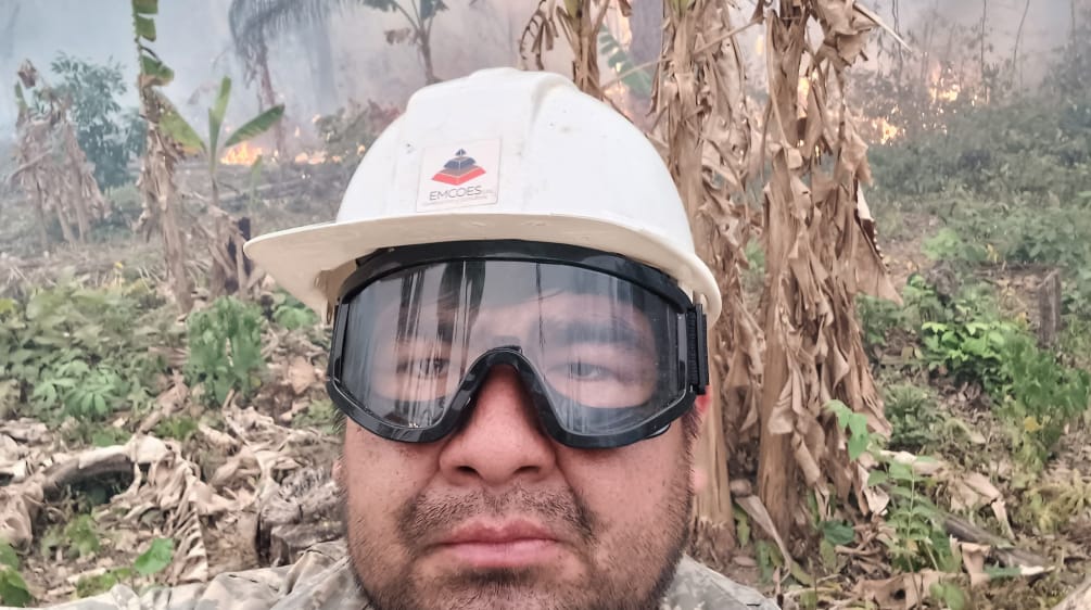 Ein Mann blickt mit einem Helm und einer Schutzbrille in die Kamera, während im Hintergrund Büsche brennen und Rauch aufsteigt