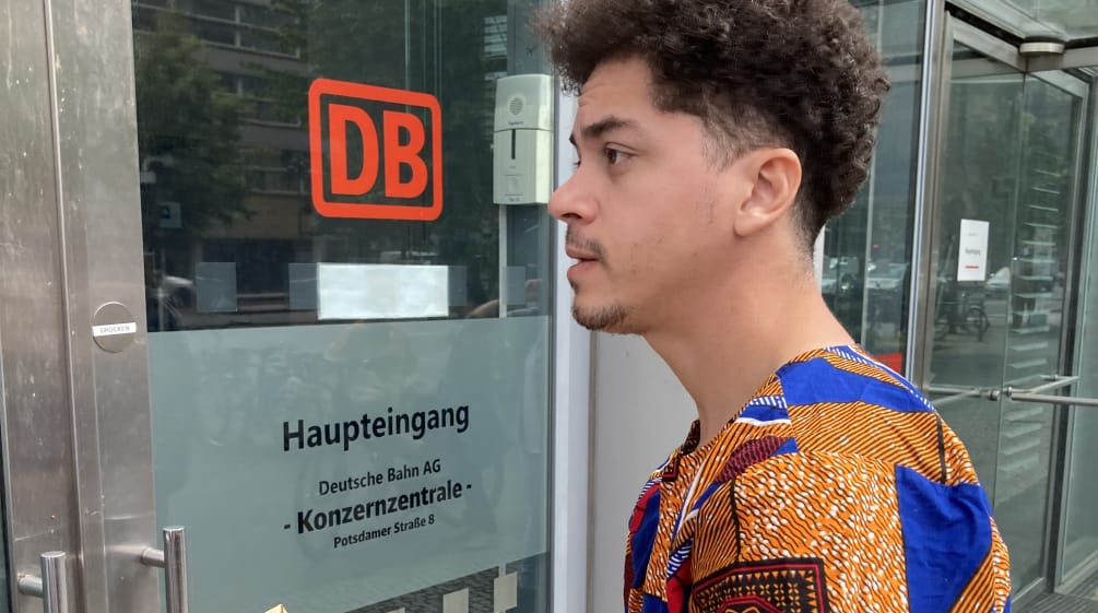 Ein junger Mann steht mit einem Briefumschlag vor einer Glastür mit der Aufschrift DB Haupteingang Konzernzentrale
