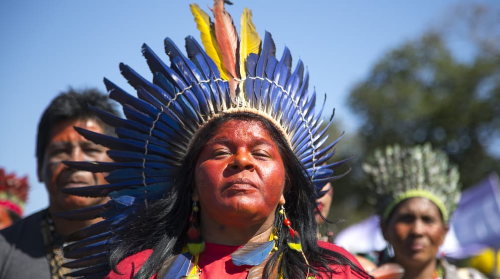 Mit einer Federkrone geschmückte indigene Frau vor zwei weiteren indigenen Personen